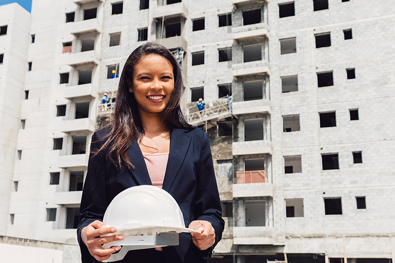 Mulher com capacete branco de engenheiro na mão e sorrindo.